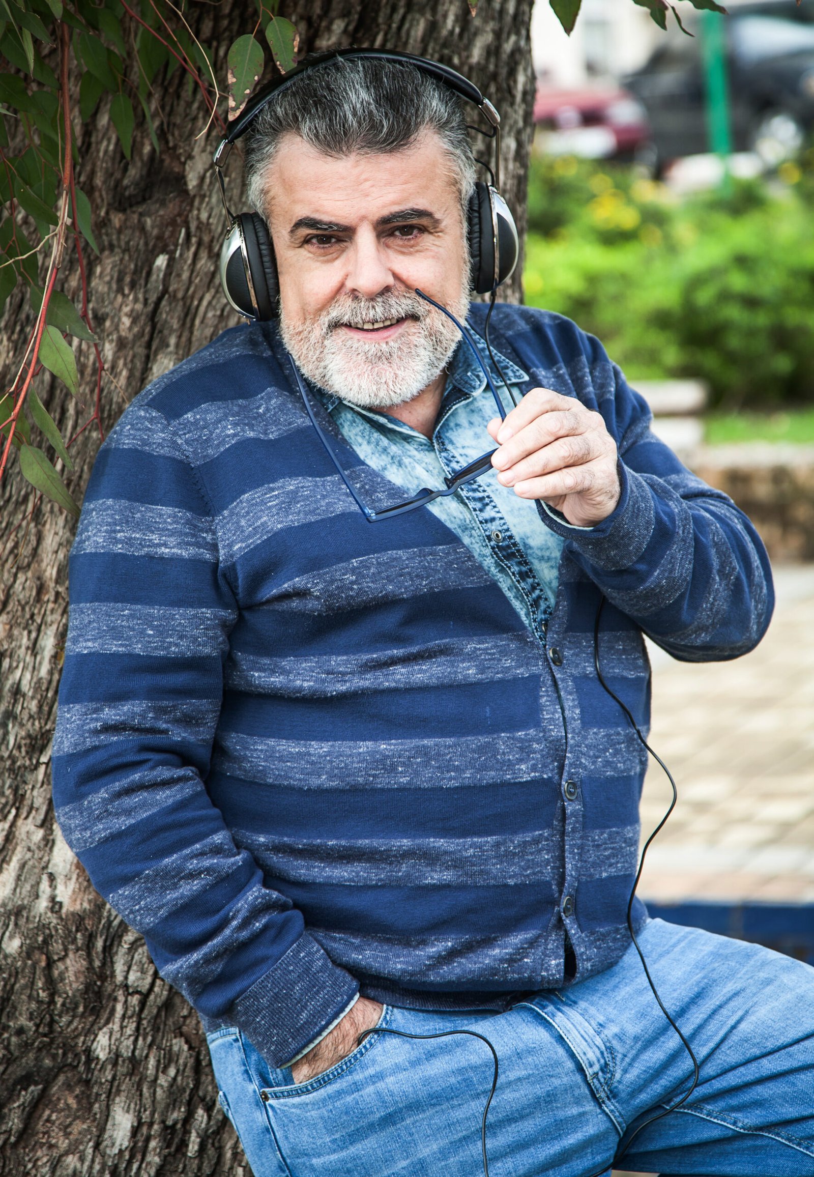 portrait smiling mature man holding sunglasses listening music while standing against tree trunk scaled